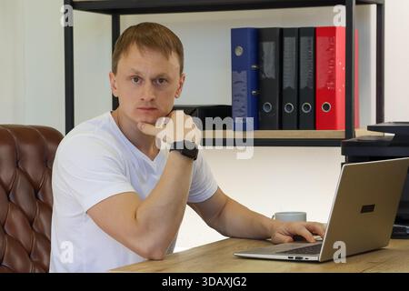 Young adult male in casual attire contemplating with hand on chin beside laptop in office setting featuring binders and office supplies. Stock Photo