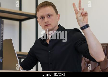 Attentive young man at desk with laptop displays victory sign or two fingers up in a professional workspace. Stock Photo