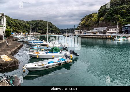 Several fishing boats lie moored close together in the harbour of Taiji, Wakayama Prefecture, Japan. Wooded hills and traditional houses line the calm water. Stock Photo