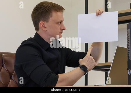 Profile view of young adult male in office, extending arm with thumbs up towards held blank white paper. Stock Photo