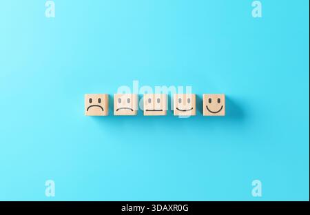 Customer satisfaction and feedback concept with wooden blocks displaying different emoji faces from sad to happy on blue background, representing user Stock Photo