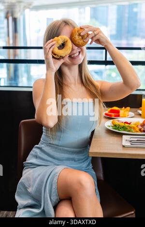 Woman in cafe playfully holding bagels over eyes, smiling, casual Stock Photo