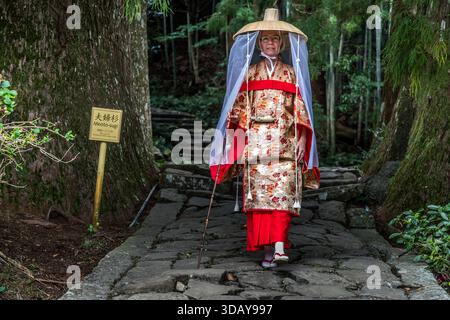 Dressed in a kimono with white socks, wooden clogs and a hat with a veil to protect against insects, the kimono experience includes walking along the old pilgrimage route, just as female pilgrims did 1,000 years ago. Person dressed in traditional Japanese clothing on the rocky Kumano Kodo pilgrimage route in Nachikatsuura. Wakayama Prefecture, Japan Stock Photo
