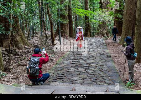 Tourist in a rented kimono on the Daimon-zaka section of the Kumano Kodo. This section of the pilgrimage route at Kumano Nachi Taisha literally means "great gate slope" and consists of 267 original stone steps. Popular photo opportunity for the Heian period attire experience. Tourists photograph a person dressed in traditional Japanese clothing on the rocky Kumano Kodo pilgrimage route in Nachikatsuura. Wakayama Prefecture, Japan Stock Photo