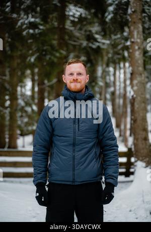 A man wearing a blue jacket stands in the snow. He is smiling and he is enjoying the winter weather Stock Photo