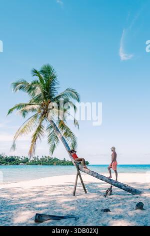 A diverse couple enjoys their time at the serene beach on Koh Kood Island, surrounded by tall palm trees and sparkling turquoise water. The warm sun enhances the tropical paradise vibe in Thailand Stock Photo