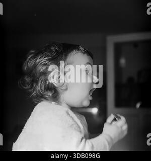 Slavyansk, Ukraine, USSR. An expressive archival portrait from the 1960s capturing a cute toddler boy indoors. The child, dressed in a soft textured shirt, is photographed in profile with his mouth wide open, yawning or singing. The dramatic side lighting creates a strong contrast against the dark background. The black and white image conveys raw emotion, innocence and the atmosphere of a happy childhood in the peaceful industrial city of Donbass during the Thaw era. Stock Photo
