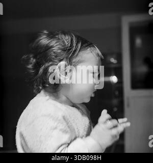 Slavyansk, Ukraine, USSR. An atmospheric archival portrait from the 1960s capturing a cute toddler boy indoors. The child, dressed in a soft textured shirt, is photographed in profile, looking to the side with a serious, focused expression and gesturing with his hands. The dramatic side lighting creates a strong contrast and highlights his hair. The black and white image conveys mood, innocence and the atmosphere of a happy childhood in the peaceful industrial city of Donbass during the Thaw era. Stock Photo
