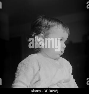Slavyansk, Ukraine, USSR. A serene archival portrait from the 1960s capturing a cute toddler boy indoors. The child, dressed in a soft textured shirt, is photographed in profile, looking away with a calm, pensive expression. The dramatic backlighting highlights his fine hair, creating a glowing effect against the dark background. The black and white image conveys innocence, peace and the artistic atmosphere of a home studio in the industrial city of Donbass during the Thaw era. Stock Photo