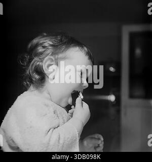 Slavyansk, Ukraine, USSR. A candid archival portrait from the 1960s capturing a toddler boy exploring the world. The cute child, dressed in a soft textured shirt, is photographed in profile, putting a small toy or object into his mouth to taste it. The dramatic side lighting creates a strong contrast against the dark background. The black and white image conveys innocence, curiosity and the atmosphere of a happy childhood in the peaceful industrial city of Donbass during the Thaw era. Stock Photo