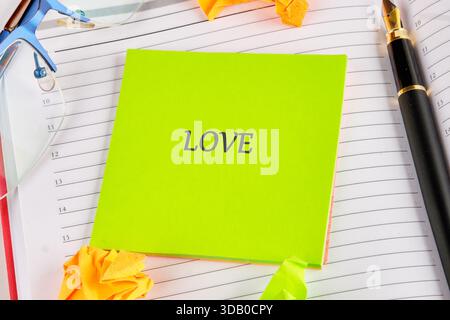 A vibrant green note shows the word love, situated on lined notebook paper. Beside it sits a black fountain pen, eyeglasses, and playful yellow origam Stock Photo