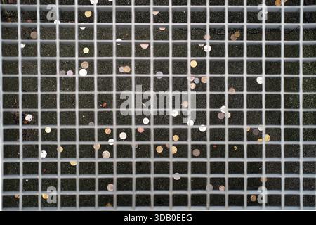 Overhead shot of galvanized steel grid flooring with rectangular mesh, coins scattered on dark stone steps below, metallic texture, neutral daylight. Stock Photo
