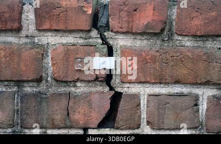 Medium view of weathered red brick wall with grey mortar and a continuous vertical crack, fitted with a transparent crack gauge at mid-height, Stock Photo