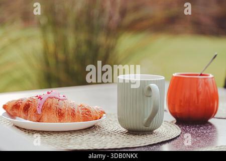 A flaky butter croissant with pink icing rests on a white plate beside a pale green mug and an orange sugar bowl outdoors. Soft natural light creates Stock Photo