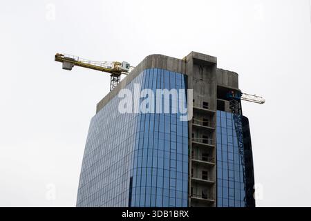 glass tower under construction with crane, blue reflective facade and exposed concrete core, rooftop crane poised Stock Photo