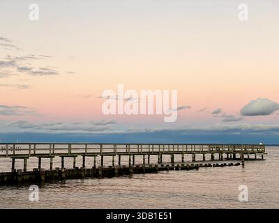 Wooden pier extending over calm water at sunset beneath a pastel-colored sky Stock Photo