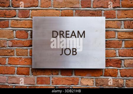 A silver sign on a brick wall showcases the words DREAM JOB in simple letters. The aged brick and bright sign create a contrast, signifying hope. Stock Photo