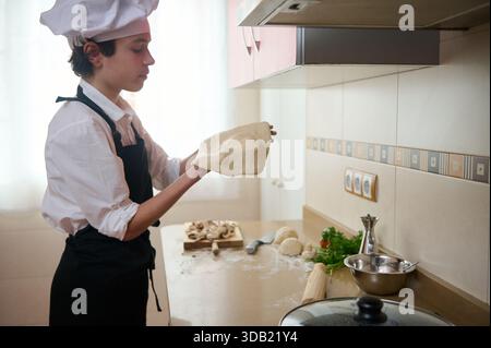 A focused young chef in a white shirt, apron, and hat kneads dough, preparing pizza dough with herbs on a clean kitchen counter. Baking tools, vegetab Stock Photo
