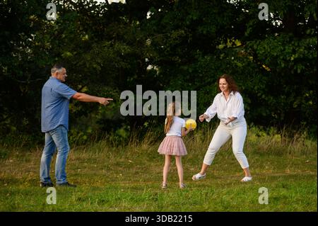 Joyful family time outdoors. Parents and daughter happily playing outside. Mom, dad, and daughter play together with ball on lawn, throwing it to each other. Summer outdoor activities Stock Photo