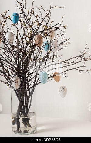 Decorative spring branches in glass vase decorated with hanging Easter eggs. Minimal Easter decor concept with natural elements and white background. Stock Photo