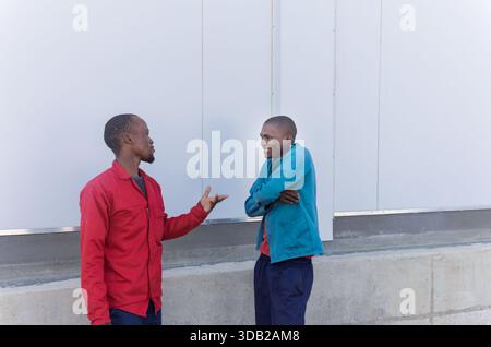 two african young workers, workwear uniform, chatting outdoors next to a warehouse wall Stock Photo