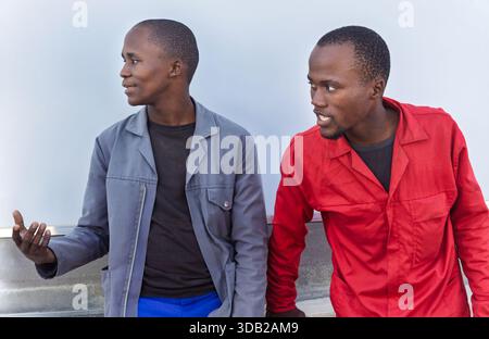 two african young workers, workwear uniform, having a talk, standing outdoors next to a warehouse wall, resting on the lunch break Stock Photo