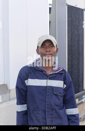 single african young worker, workwear uniform silver reflective safety stripe, with a hat, standing outdoors next to a warehouse wall Stock Photo