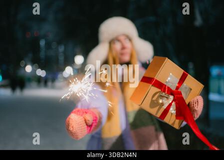 Young woman holding a glowing sparkler and a festive gift package, enjoying the joyous atmosphere of winter holidays and special celebrations in the evening snow Stock Photo