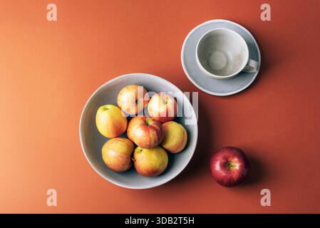 Fresh ripe apples in a bowl and an empty cup with saucer on an orange background. Top view, flat lay, copy space. Stock Photo
