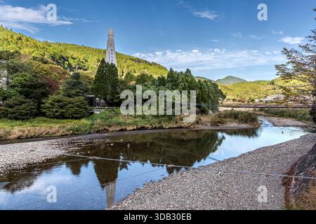 A tall obelisk inscribed with the characters for "Watarase" stands on the bank of the Yomura River in the Hongu Valley, near Kumano Hongū Taisha. The forested mountains are reflected in the calm water. Hongucho Wataze, Tanabe, Wakayama Prefecture, Japan. Stock Photo