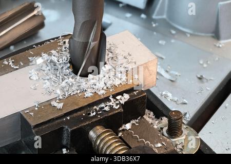 Solid core drill bit enlarge hole in aluminum profile and metal chips. Closeup sharp steel cutting tool at working in workpiece clamped in bench vise. Stock Photo