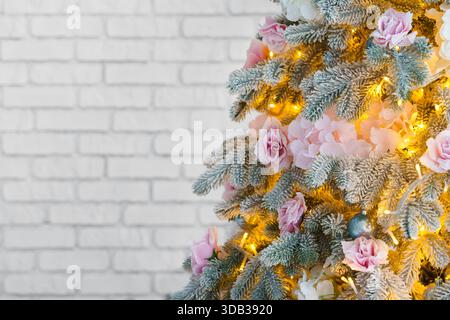 Close up of frosted, snowy Christmas tree decorated with pink roses, white flowers, and warm glowing lights on white brick wall background with copy s Stock Photo