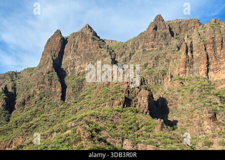 Imposing rugged mountain cliffs densely covered in sparse green vegetation. Steep, rocky slopes dominate the frame against a partly cloudy blue sky. I Stock Photo