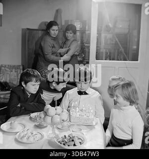 Sloviansk, Ukraine, USSR. February 5, 1977. A black and white archival photograph captures a heartfelt birthday celebration in a Soviet apartment. A boy in a white shirt, the birthday boy, sits at a festive table with a homemade cake and lit candles, surrounded by friends. His mother and sister stand in the background, watching warmly. The table is set with apples, sweets, and tea cups on a white cloth, reflecting the modest yet cozy atmosphere of a peaceful childhood in the Donbass region during the 1970s. Stock Photo