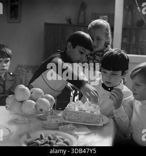Sloviansk, Ukraine, USSR. February 5, 1977. A dynamic black and white archival photograph captures a collaborative moment at a birthday party. A friend in a dark shirt leans over the table to help adjust candles on the homemade cake, while the birthday boy in a white shirt and bow tie watches closely. Surrounded by other friends, the group anticipates the celebration. The table features apples and sweets, reflecting the camaraderie and warmth of a peaceful childhood in the Donbass region during the 1970s. Stock Photo