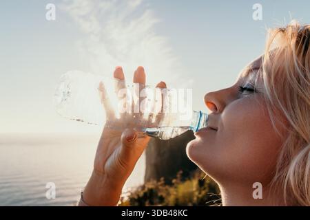 Woman Water Bottle Ocean Sunset - Young woman drinks water from a bottle while looking at the sunset over the ocean. Stock Photo