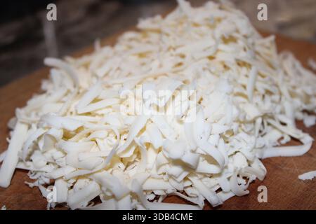 Shredded cheese sits on a wooden cutting board in a kitchen. It is prepared for use in various dishes. The light in the room is dim as evening approac Stock Photo