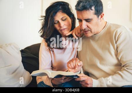Middle-aged couple sitting on a beige sofa at home reading a book. Man pointing at a specific passage while woman holds the book. Authentic lifestyle Stock Photo