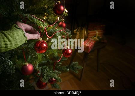 Hand hanging red baubles and decorations on the Christmas tree, preparing for the holiday at home, dark background, copy space, selected focus, narrow Stock Photo