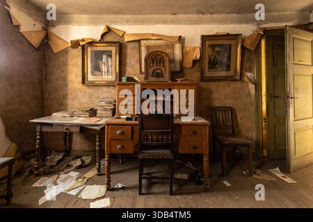 Abandoned vintage study with wooden desk, antique radio, peeling wallpaper and scattered papers in a light-filled derelict room Stock Photo