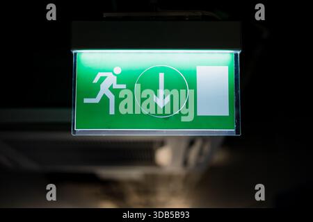 Illuminated green emergency exit sign with running figure and downward arrow mounted indoors showing safety guidance and evacuation direction Stock Photo