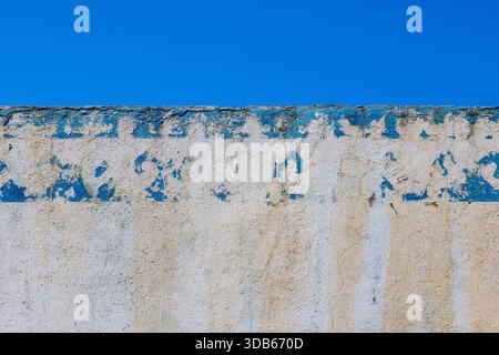 Textured wall features peeling blue and beige paint in outdoor setting. Bright sky enhances rustic mood, showcasing age and character of the structure. Stock Photo