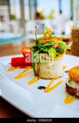 A layered salad features a croquette sandwich and red chilli peppers on a white plate. Vibrant vegetable ribbons and drizzled sauces accent the dish. Stock Photo
