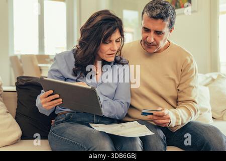 Middle-aged couple sitting on sofa managing household finances. Woman holding a digital tablet and man holding a credit card to pay bills online. Pape Stock Photo