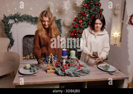 Two women setting festive Christmas table with evergreen wreath centerpiece, candles and wine glasses in cozy living room. Stock Photo