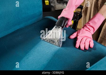 Female worker in rubber gloves wet vacuums blue upholstered chair seat during professional home stain removal service Stock Photo