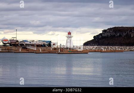 White and red wooden lighthouse beside drydocked boats on a rocky shoreline with rugged cliffs at Lion’s Head, Canada, under a winter sky. Stock Photo