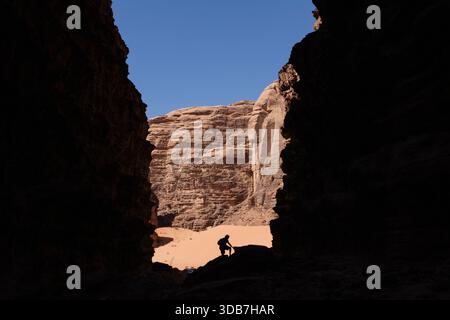 A person appears in silhouette inside a canyon in Wadi Rum, Jordan, surrounded by high sandstone walls and bright sunlight illuminating the rocks beyo Stock Photo
