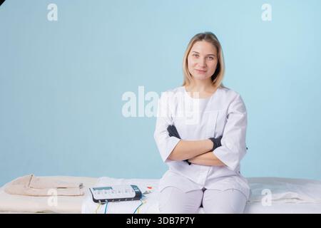 Confident female therapist in a white uniform with black gloves sitting in a wellness clinic, ready for a treatment session with electrotherapy equipm Stock Photo