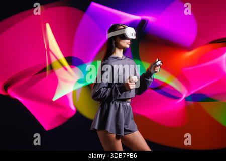 Young woman in VR headset playing virtual reality game with motion controllers, neon light trails in futuristic immersive environment. Stock Photo